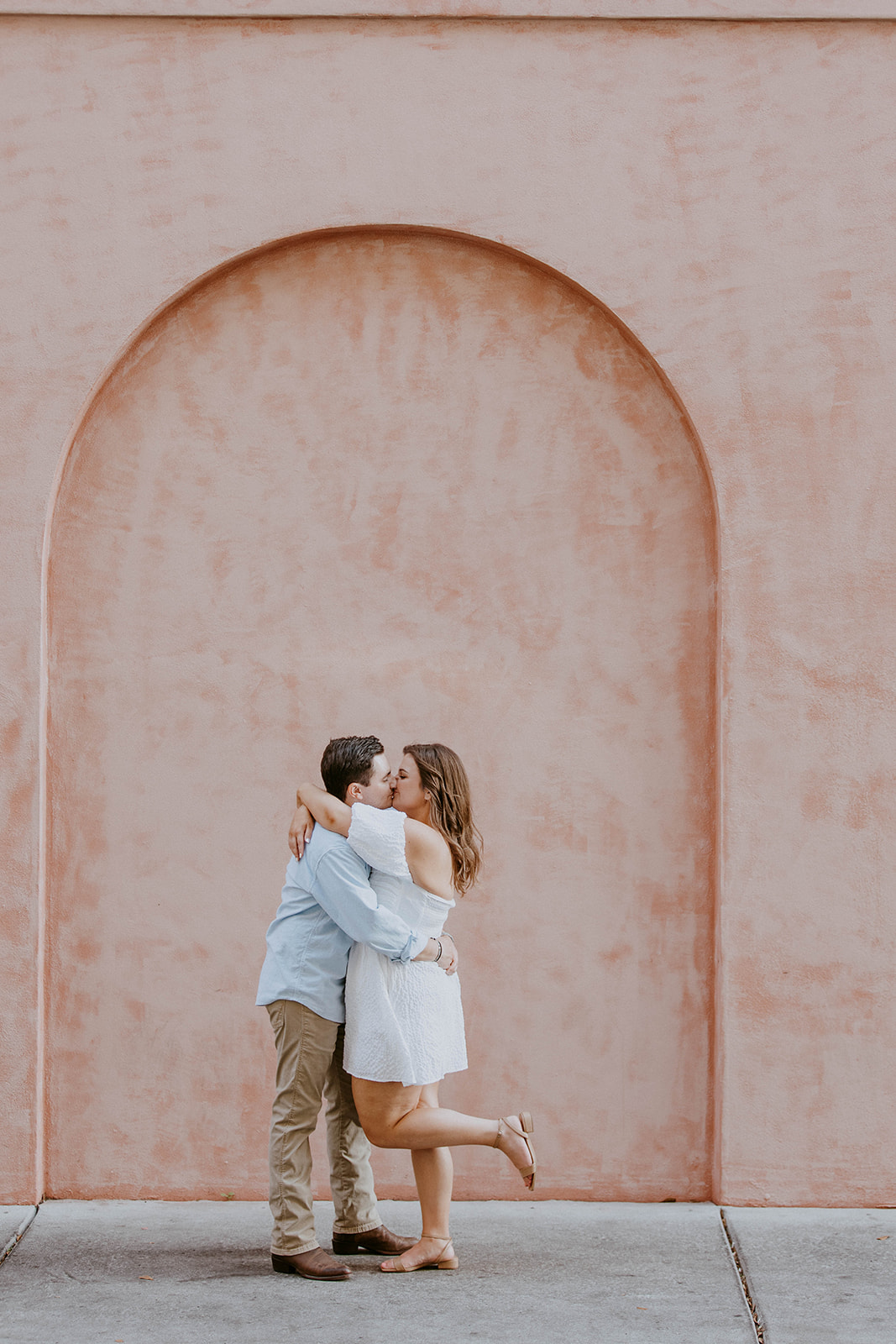 A couple stands holding hands in front of a pink wall. The man is wearing a light blue shirt and khaki pants; the woman is in a white off-shoulder dress. They are both looking in opposite directions.