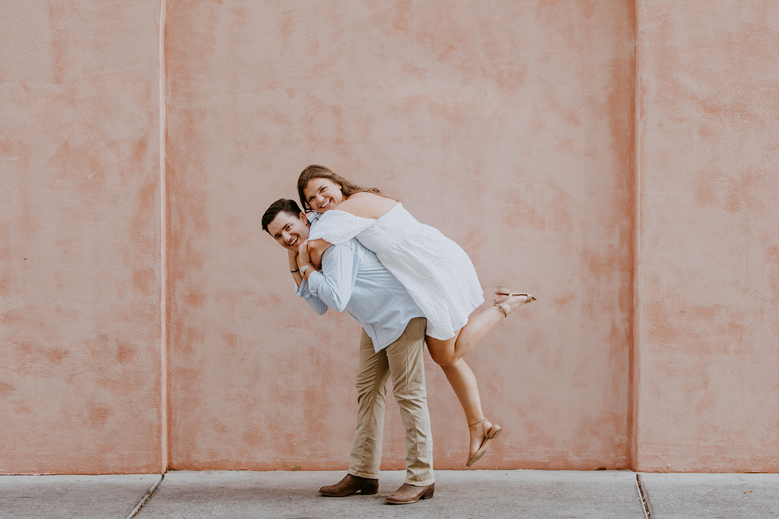 A couple stands holding hands in front of a pink wall. The man is wearing a light blue shirt and khaki pants; the woman is in a white off-shoulder dress. They are both looking in opposite directions.