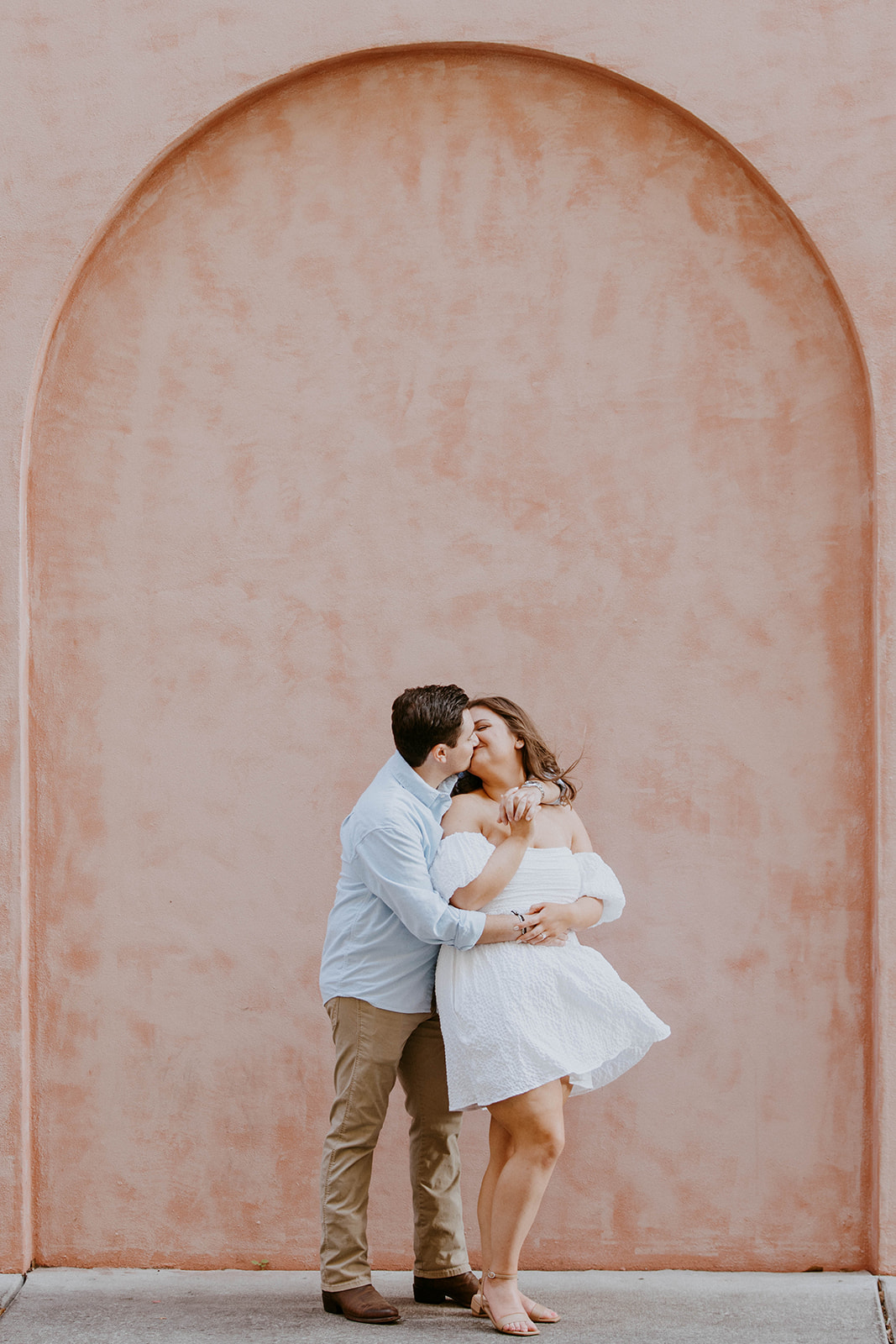 A couple stands holding hands in front of a pink wall. The man is wearing a light blue shirt and khaki pants; the woman is in a white off-shoulder dress. They are both looking in opposite directions.