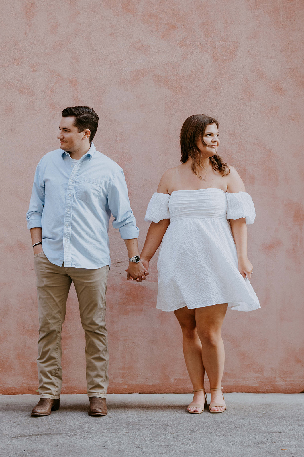 A couple stands holding hands in front of a pink wall. The man is wearing a light blue shirt and khaki pants; the woman is in a white off-shoulder dress. They are both looking in opposite directions.