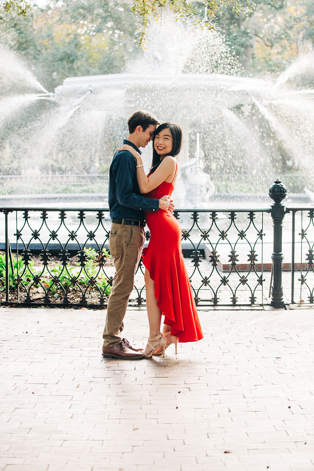 A couple is kissing on a sidewalk near a black iron fence. The man is wearing a blue shirt, and the woman is in a red dress at forysth park in savannah for their engagement