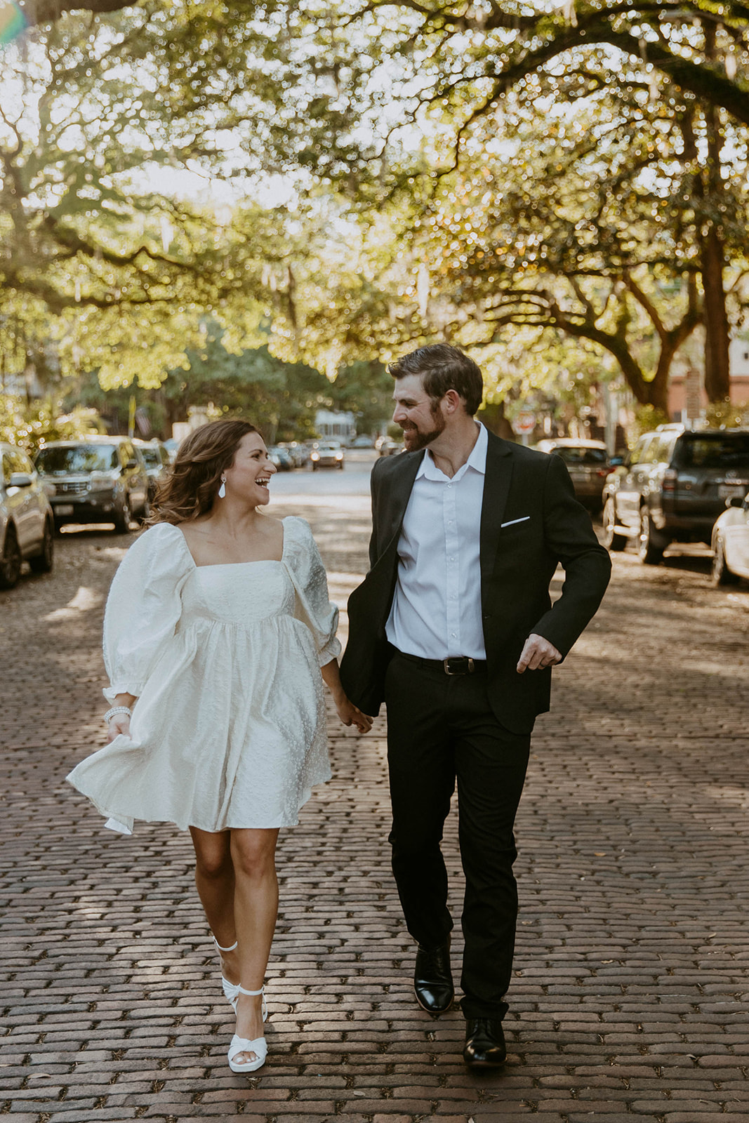 Two people, one in a white dress and the other in a black suit, hold hands while walking on a tree-lined cobblestone street on Jones street fir their savannah engagement 