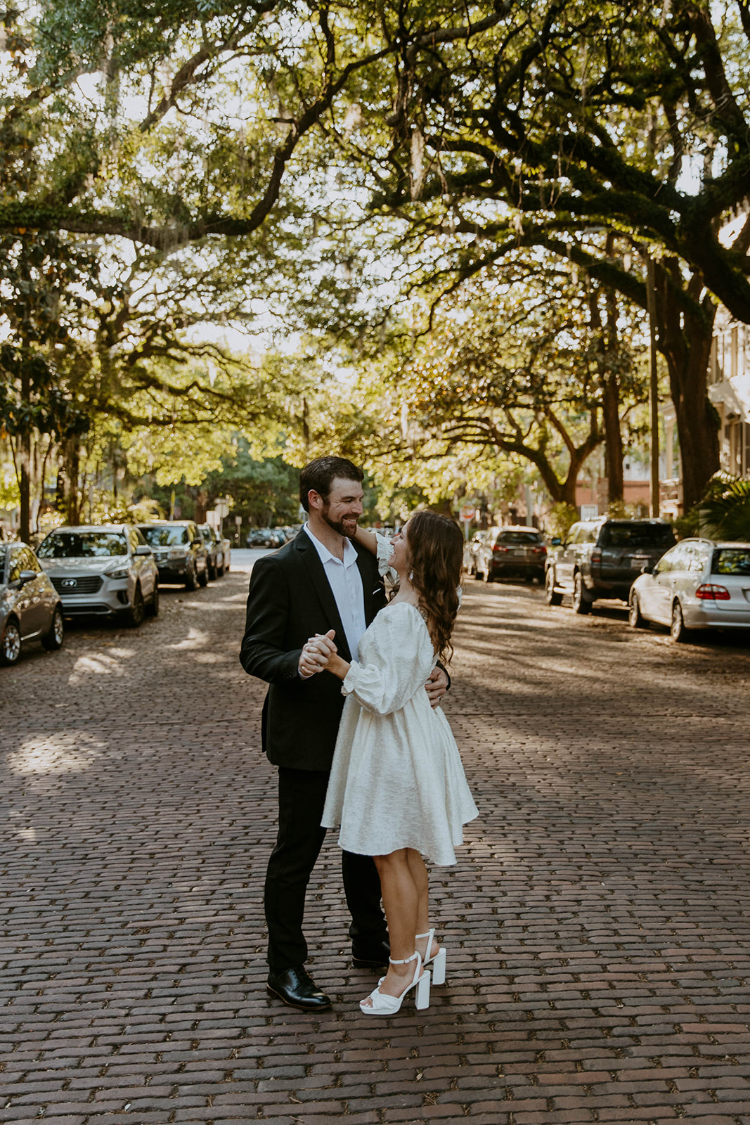 Two people, one in a white dress and the other in a black suit, hold hands while walking on a tree-lined cobblestone street on Jones street fir their savannah engagement 