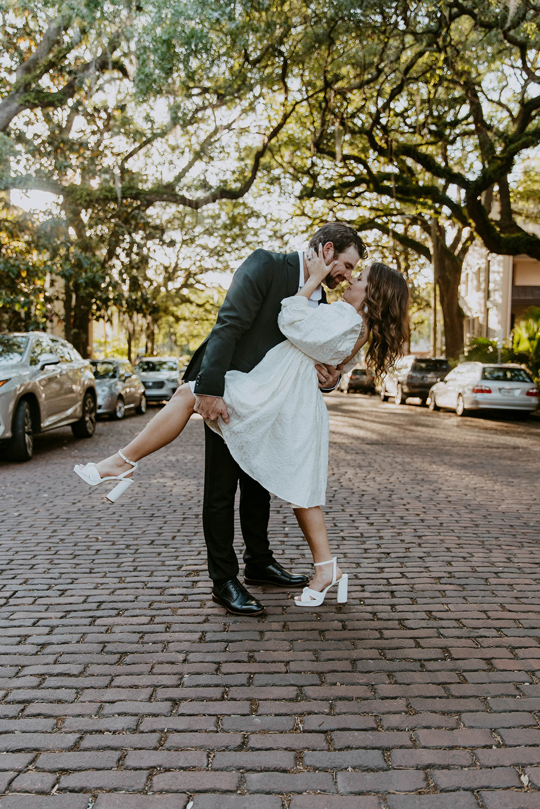 Two people, one in a white dress and the other in a black suit, hold hands while walking on a tree-lined cobblestone street on Jones street fir their savannah engagement 