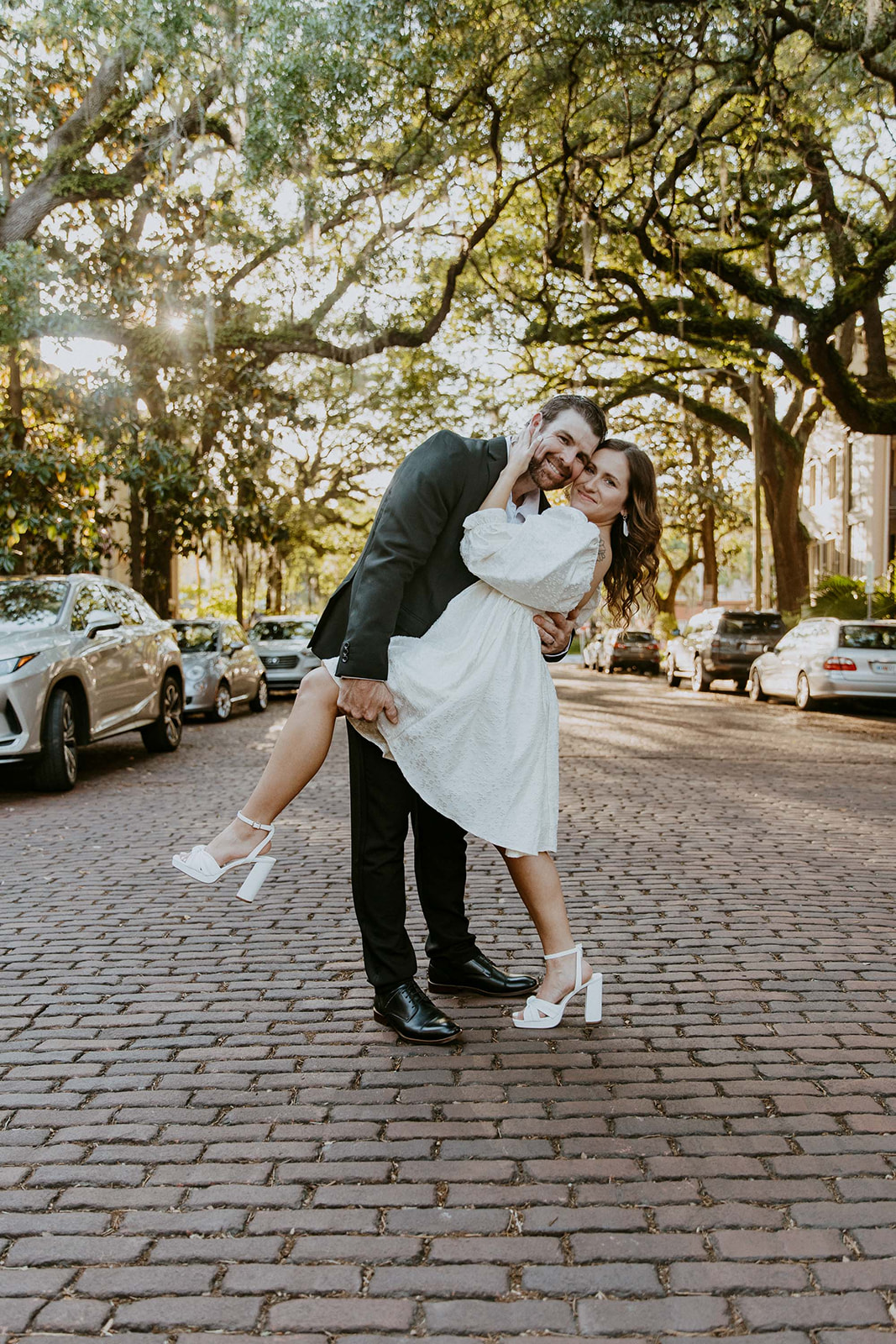 Two people, one in a white dress and the other in a black suit, hold hands while walking on a tree-lined cobblestone street on Jones street fir their savannah engagement 