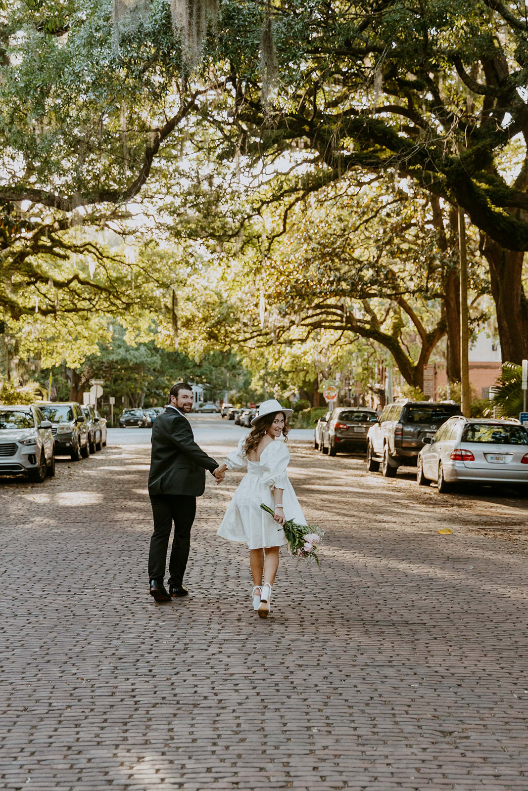 Two people, one in a white dress and the other in a black suit, hold hands while walking on a tree-lined cobblestone street on Jones street fir their savannah engagement 