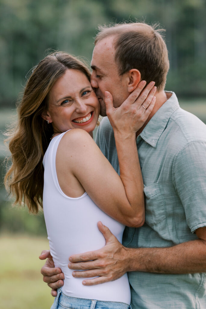 A man and woman embracing, he is kissing her on the cheek while she is smiling and looking towards the camera. 