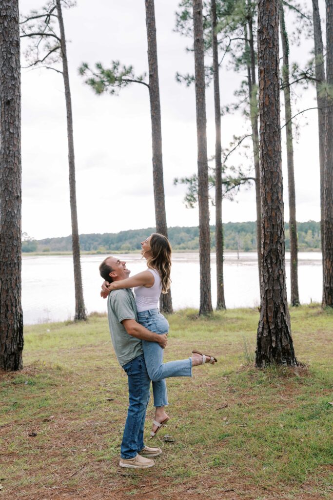 Man and woman embracing in the woods, he is lifting her off the ground while they are both smiling. 