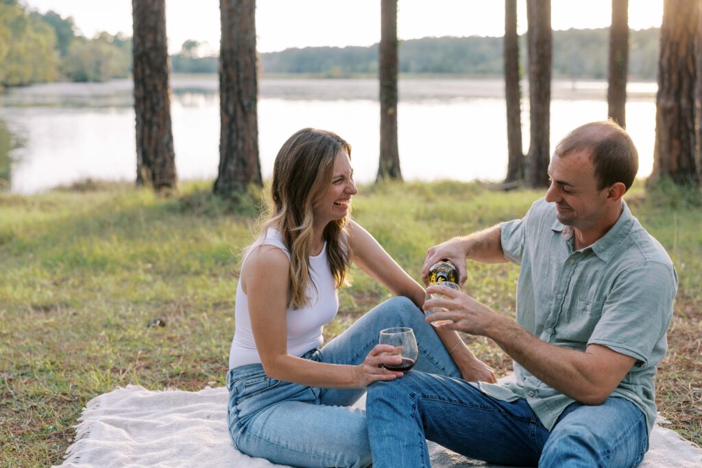 A man and woman sitting on a blanket in the woods, while he is pouring a glass of wine and she is laughing. 