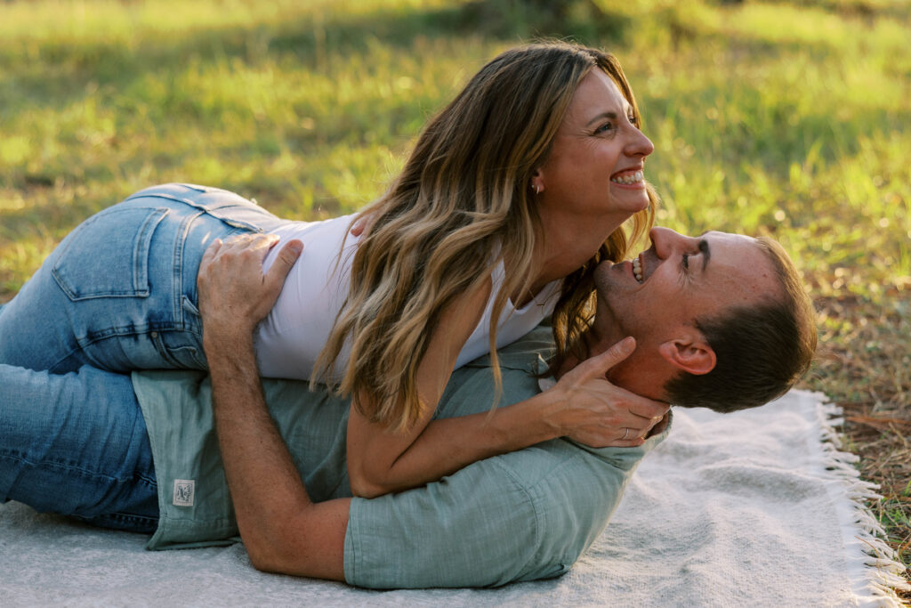 A man and woman laying on the ground on a woven blanket, they are laughing. 