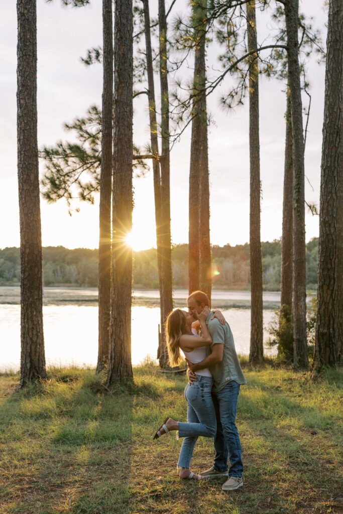 A man and woman embracing and kissing in the woods while sun is coming through the trees. 