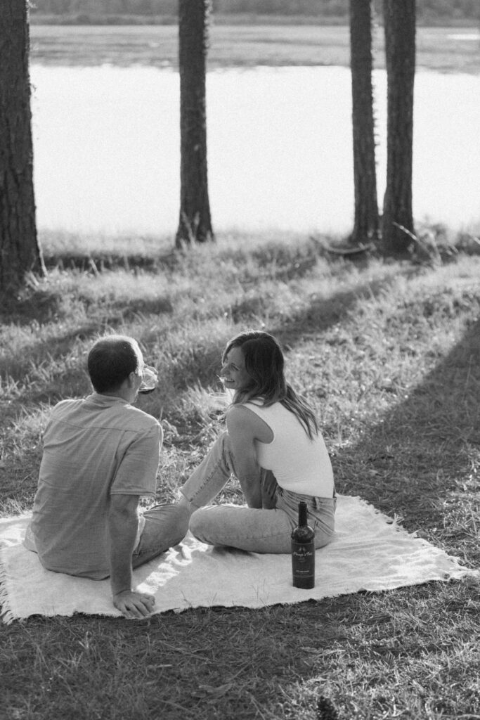 A man and woman sitting on a blanket in the woods, they are talking and enjoying a glass of wine